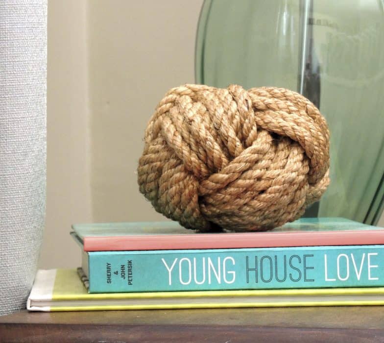 A large ball of rope, resting on a stack of books on an end table, in front of a lamp with a green, glass base