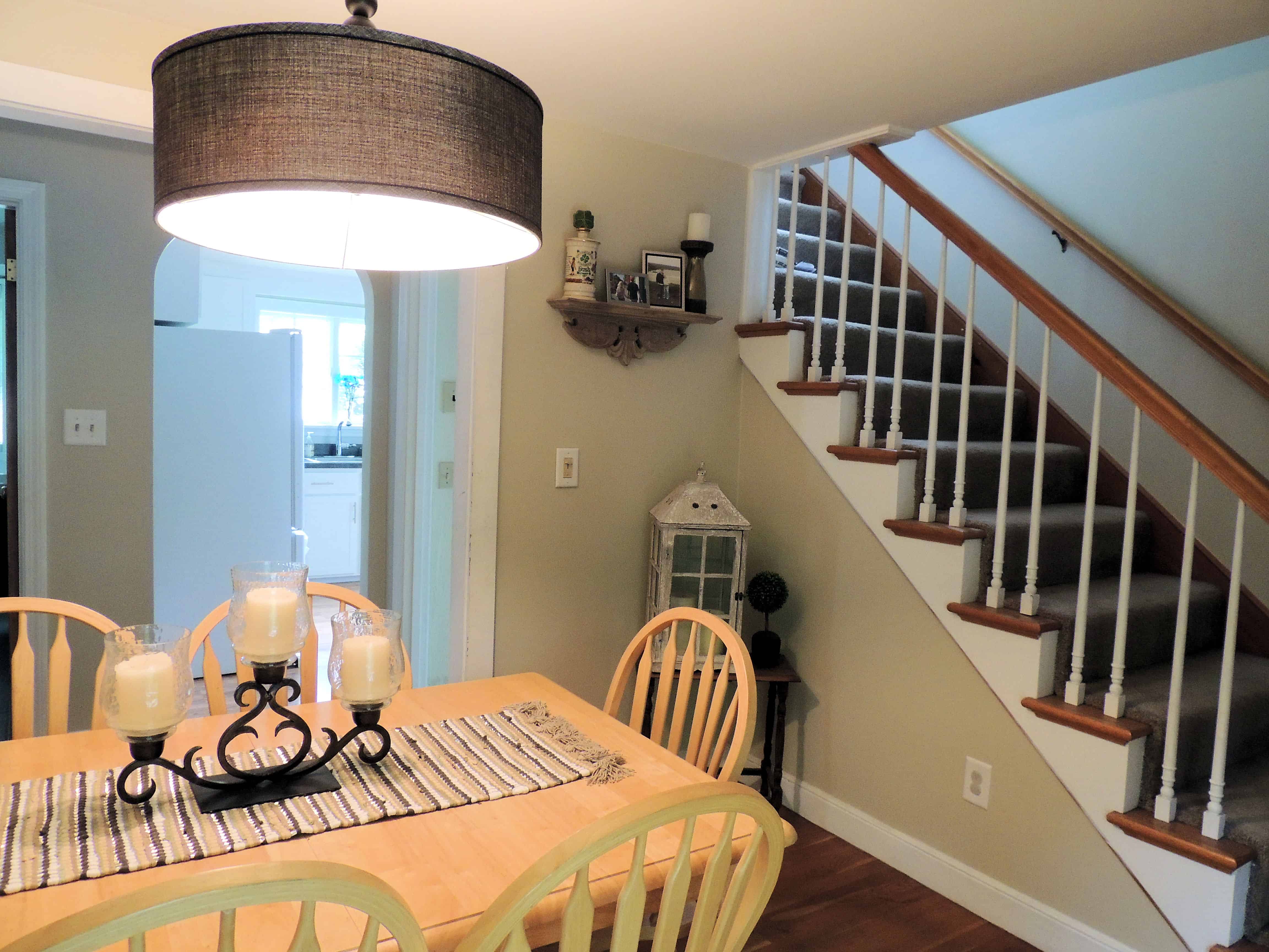 A pale, wooden table with matching chairs in a dining room with a large, drum-shaped light fixture overhead, and a staircase in the background