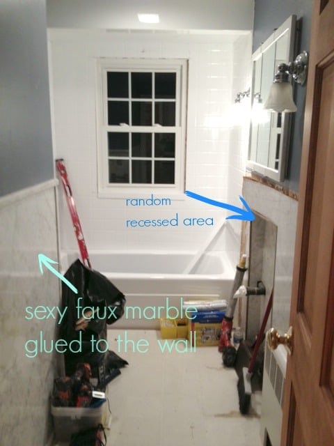 A bathroom with faux marble glued halfway up the wall on the left, a recessed area on the right below a mirror and a window and bath tub in the background