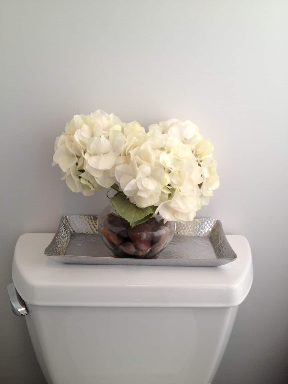 A vase of large, white flowers resting on a rectangular, silver tray on the tank of the toilet
