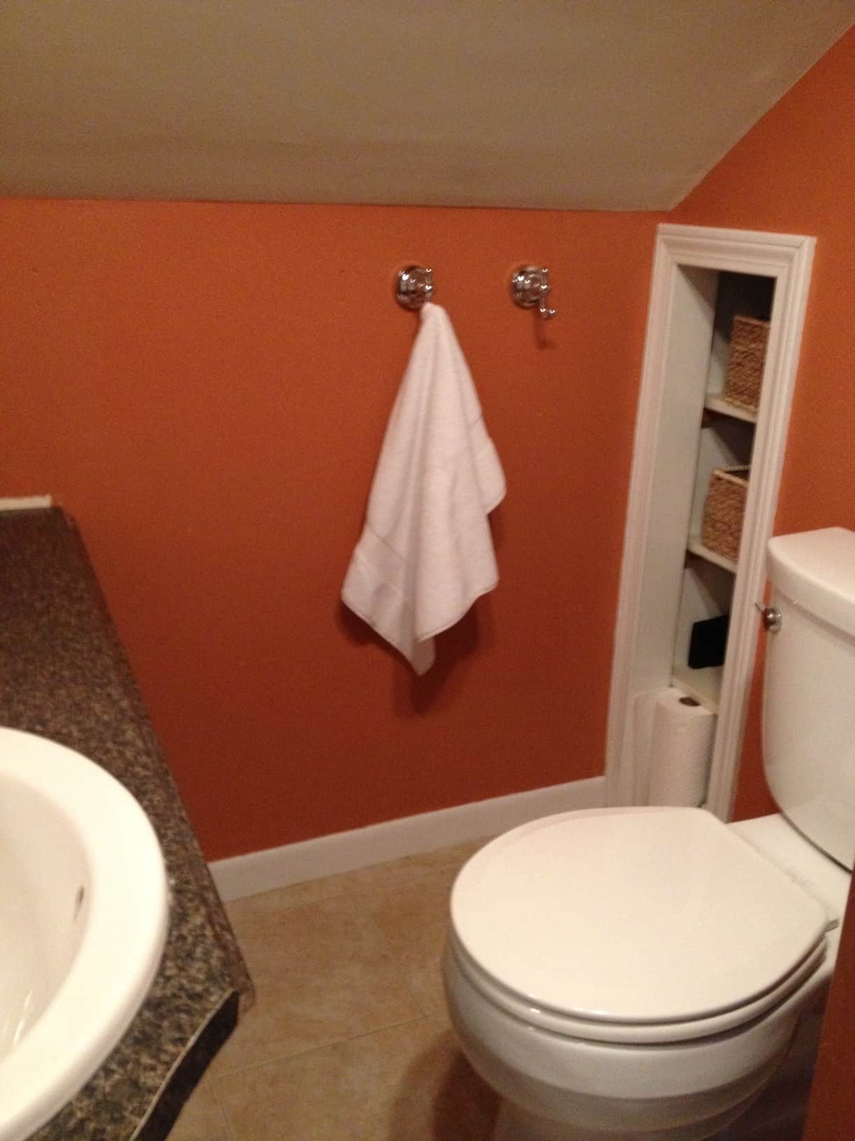 A bathroom with orange painted walls, silver hooks for towels, a recessed shelf for supplies and a sink and toilet nearby
