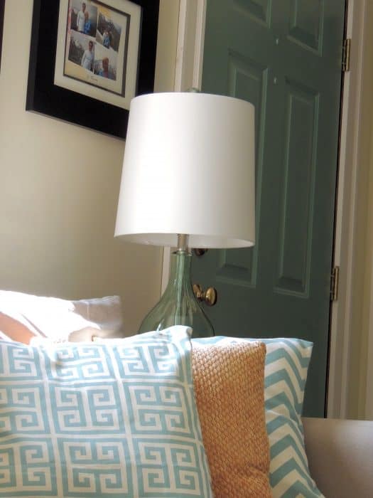 A trio of pillows in tan, blue and white, resting on a sofa, with a lamp with a green, glass base in the background