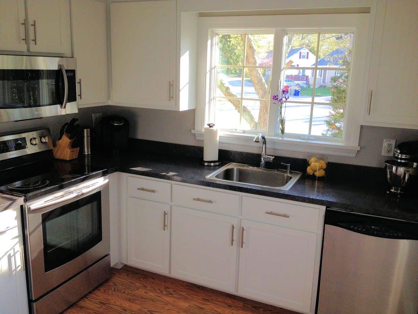 A kitchen with a large window above a kitchen sink, white cabinets and dark countertops