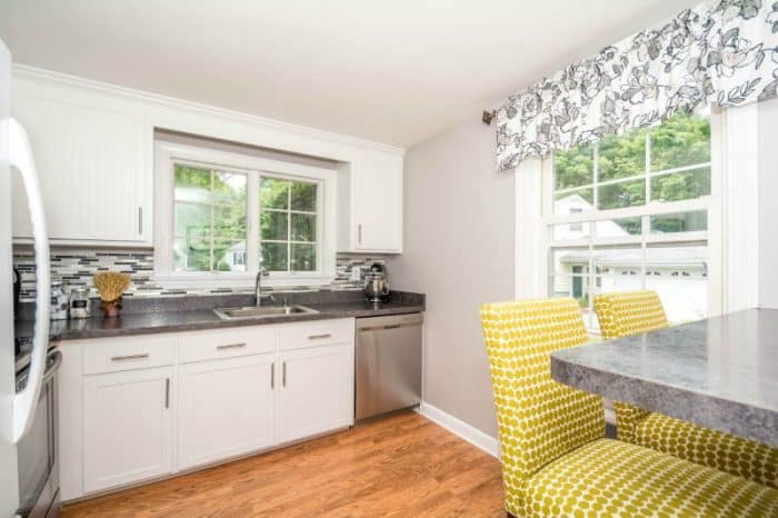 The corner of a kitchen with white cabinets, wood floors, and two large windows nearby