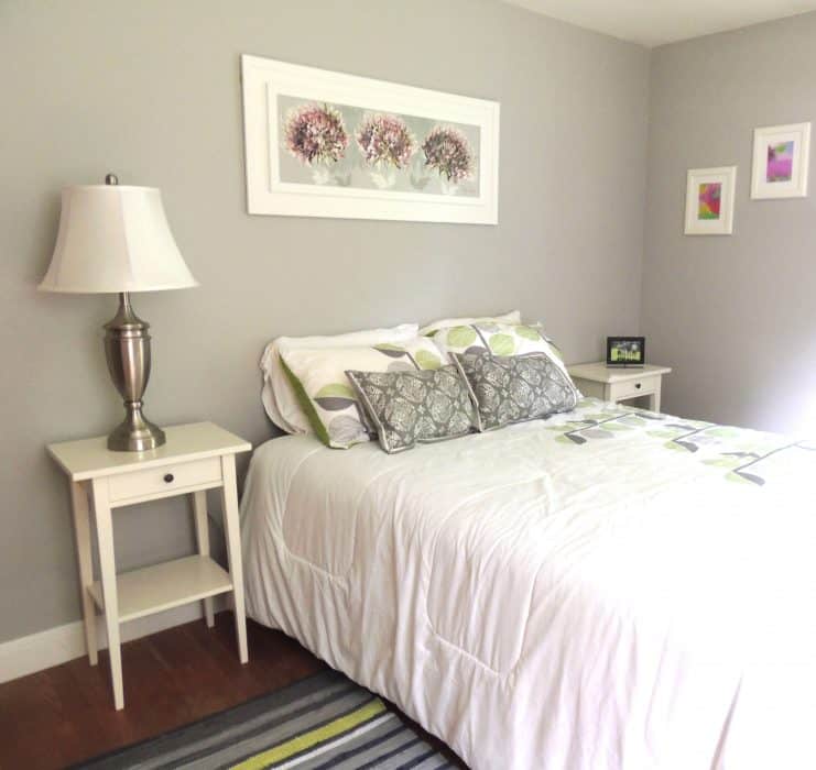 A bed with grey and white bedding, on wood floors with a striped rug, and various artwork on the walls 