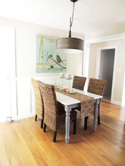 A dining room with a table, wicker chairs, wood floors and a large, square  painting on the wall