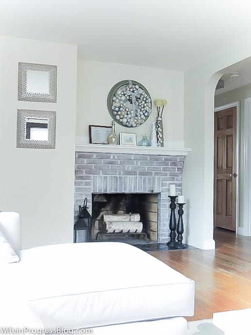 A whitewashed fireplace with white wooden mantel holding a vase, frame and other blue and white decor, with the wreath hanging above on the wall