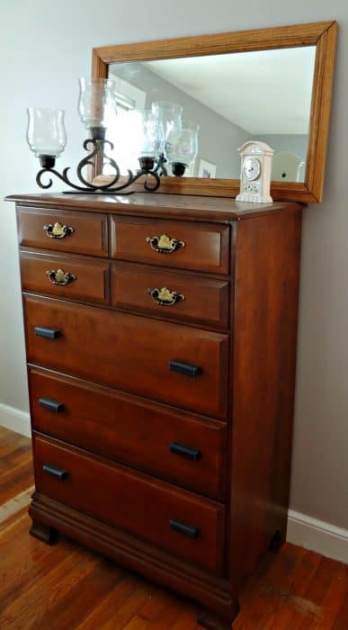 A wooden chest of drawers with a large mirror, metal candle holders and a small, beige clock