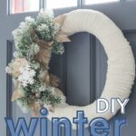 A handmade winter wreath using yarn and artificial flowers hanging on a blue door.