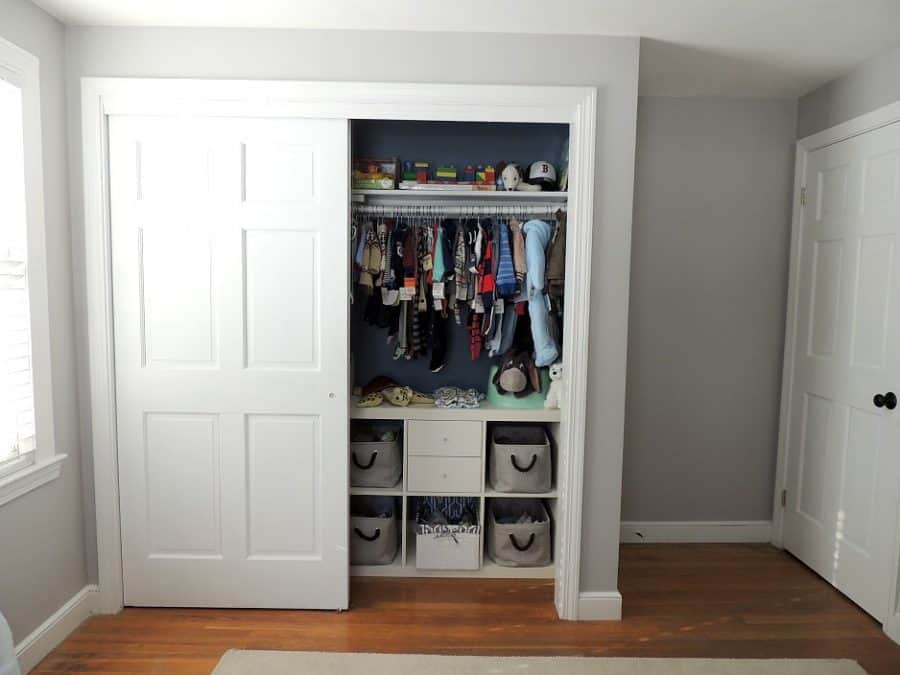 A closet with one door open to reveal baby clothes hanging from a rack, a shelf above holding caps and a shelving system below holding grey baskets