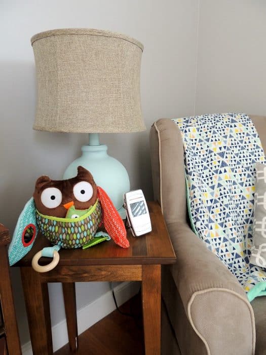 A grey, upholstered rocking chair next to dark, wooden side table holding a lamp with a blue base and beige shade, and a stuffed owl toy