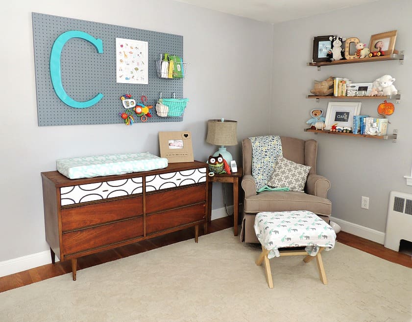 A bedroom with a dark brown dresser/changing table combo, a grey pegboard with a large initial \'C\' in blue and a brown rocking chair nearby