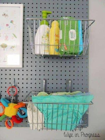 A large, grey pegboard with silver, wire baskets holding baby bath supplies and diapers