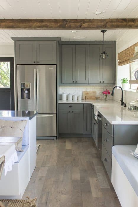 A kitchen with grey cabinets, stainless steel appliances, farmhouse sink, grey wood floors, and a painted wood plank ceiling with light brown cross beams