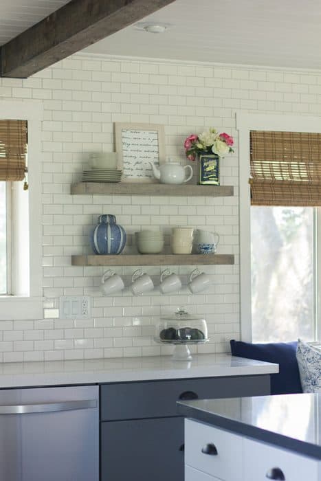 A kitchen wall with two floating wooden shelves with miscellaneous decor on top and hooks with mugs hanging underneath
