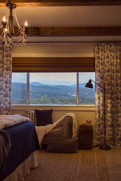 In the foreground, the edge of a bed and in the background, a large, wide window showing a mountain scenery, with floor-to-ceiling curtains and a sofa below the window