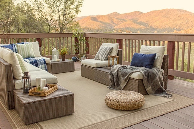 A deck with grey, wicker seating, white cushions and blue pillows & throws, on a tan outdoor mat, with the mountains in the distance