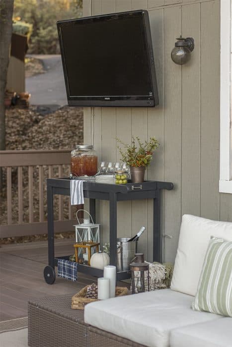 The corner of a deck where a television is mounted from the side of the house, above a grey beverage cart containing drinks, glassware and a vase of flowers