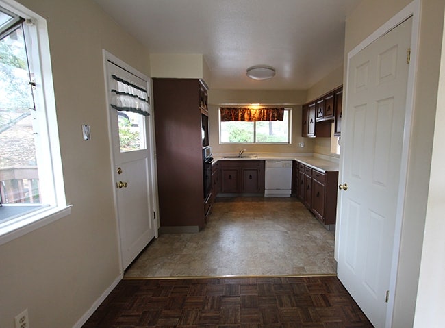 A kitchen with dark brown cabinets, tiled floors, short curtains above window and white appliances