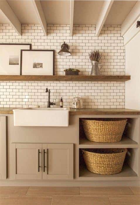 A kitchen with small, subway tile wall, a floating wooden shelf with framed art and other decor, a farmhouse sink, and grey cabinet with shelves below for wicker baskets