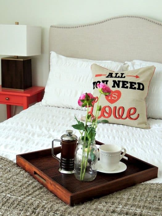 A rectangular, wooden tray holding a tea cup, saucer, glass vase with flowers and a coffee press on a bed 