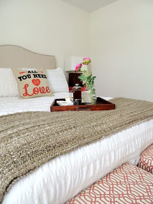 A bed with white comforter, accent pillow, tray of coffee and two ottomans at the end with a red and cream pattern