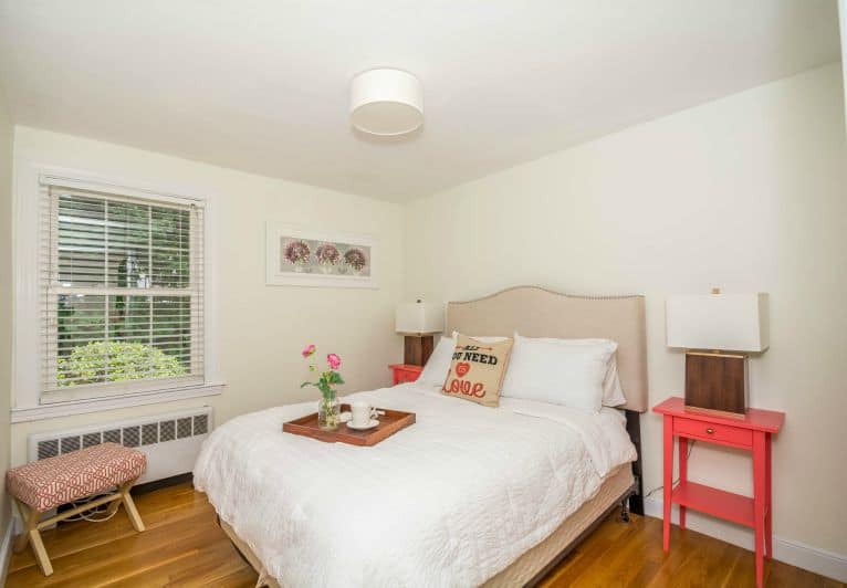 A bed with a beige headboard and white bedding, holding a wooden tray and vase of flowers, with a bright, pink night stand nearby