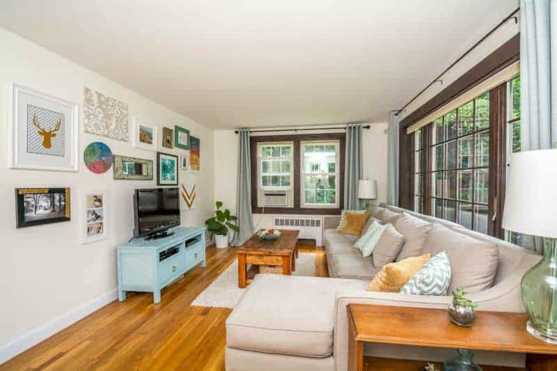 A living room, with large windows on the front and side, a large, light grey, sectional sofa, a light blue television stand and various artwork on the wall 