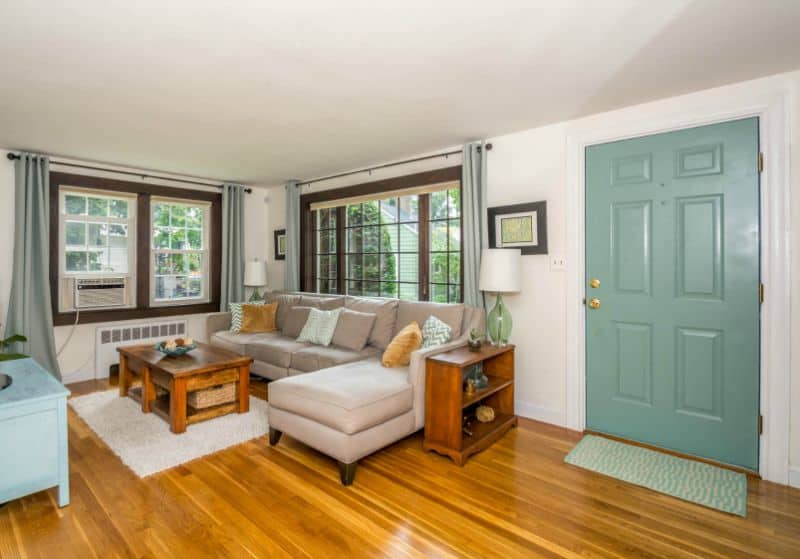 A living room with a green front door, wooden floors, two large windows, a long sofa set and wooden coffee table on a white rug