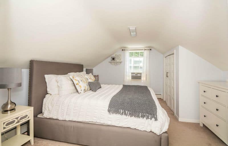 A bed with an upholstered headboard, white and grey bedding and numerous pillows, with a window in the background