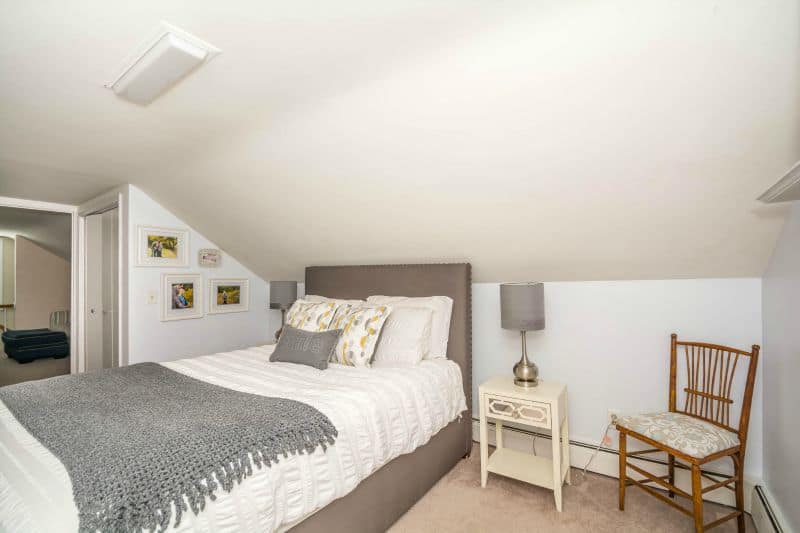 A bedroom with a grey and white bed, artwork on the wall, a beige nightstand and a lamp with a grey shade