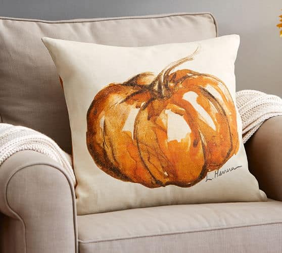 A square beige pillow with a photo of a large, orange pumpkin, resting on a light grey armchair