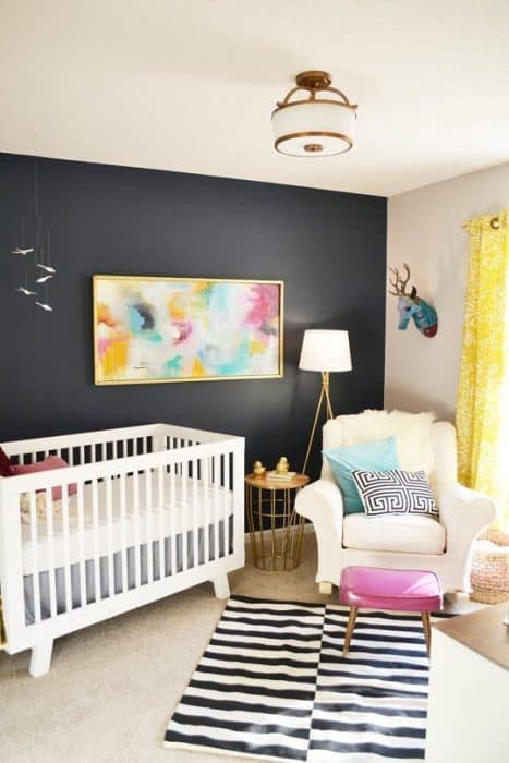 A bedroom with one navy accent wall, a colorful abstract wall painting, a white armchair, white crib, yellow curtains and a pink foot stool