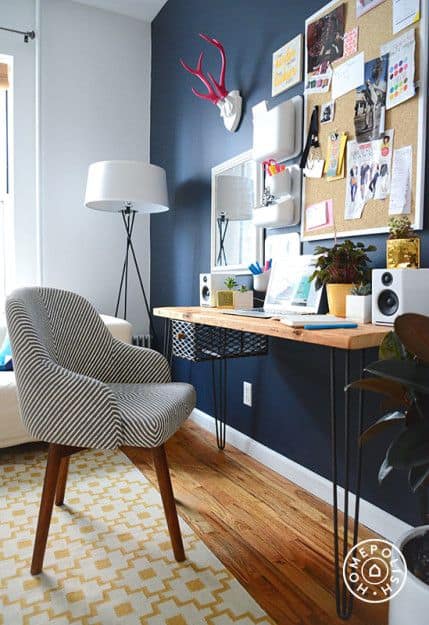 A room with a navy accent wall, mounted mirror and bulletin board and a narrow desk mounted on the wall, with an upholstered accent chair
