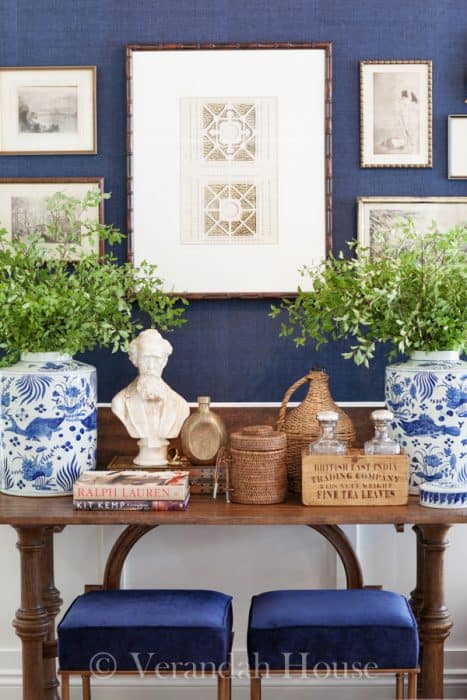 A navy wall with framed artwork of different sizes, and a side table with large blue and white ceramic vases of greenery and various wooden decor items