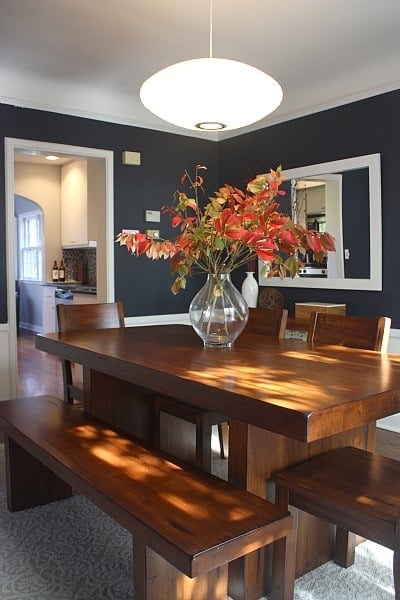 A dining room with navy walls, wood trim, and a hefty wooden table with chairs and a bench, with a large clear vase in the center, holding flowers