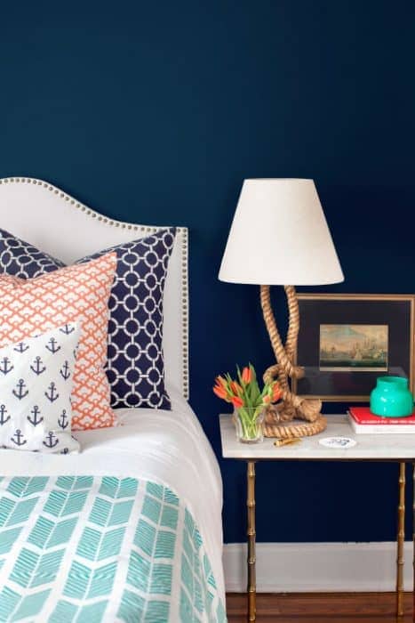 The end of a bed, with various nautical patterned pillows, a lamp shade with a rope base, and navy walls