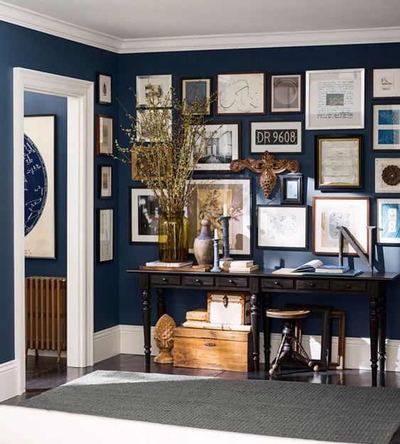 A long, narrow dark brown desk holding a green vase of greenery, resting against a navy wall, filled completely with various framed art