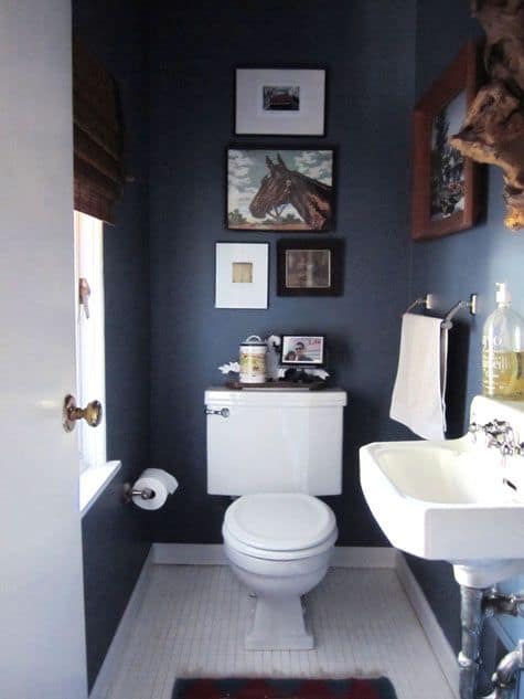 A bathroom with a window opposite the sink, and many pieces of framed art above the toilet on a dark blue wall