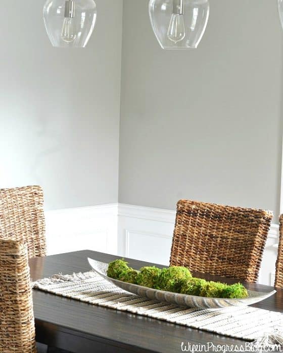 A dining room with clear, glass globe lights overhead, tall wicker chairs and a dark, wooden table with a narrow tray of moss balls