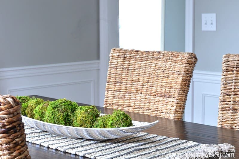 A wicker chair at a dark, wooden dining table, with a narrow, silver platter of moss balls on a thick, grey and white runner