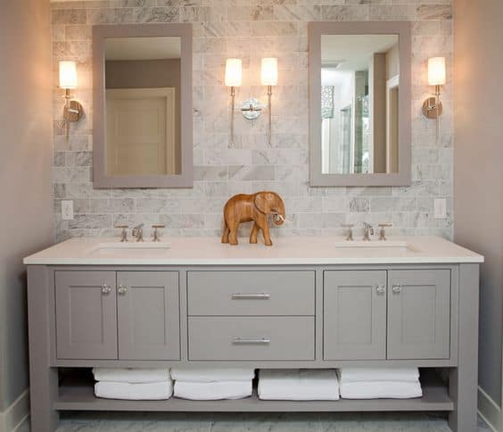 A double sink vanity with white countertop, silver faucets, silver knobs and handles on grey cabinets and a shelf below with white bath towels, and a wooden elephant on the sink top