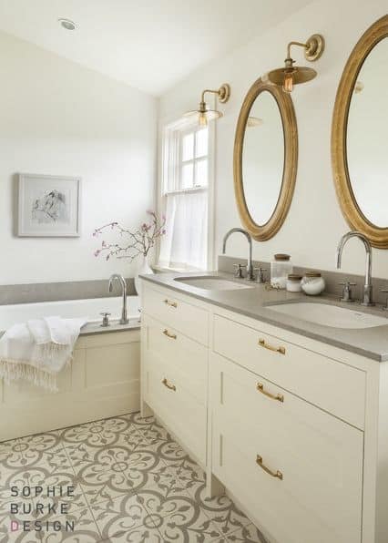 A bathroom with large bathtub with beige, wood framed side, near a beige double sink with grey countertop and oval, gold-framed mirrors 