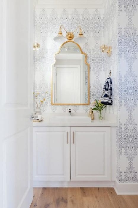 A white sink with white cabinet doors, a mirror with gold frame, and delicate blue and white wallpaper