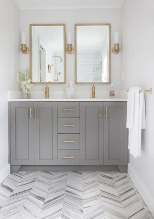 A sink with gold hardware, grey cabinets, white countertop, two mirrors with gold frames, three gold sconces and a grey, herringbone-patterned floor
