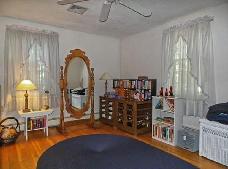 A bedroom with wood flooring and an oval, dark blue area rug, surrounded by an end table, floor-length mirror, and various shelves of books and miscellaneous items