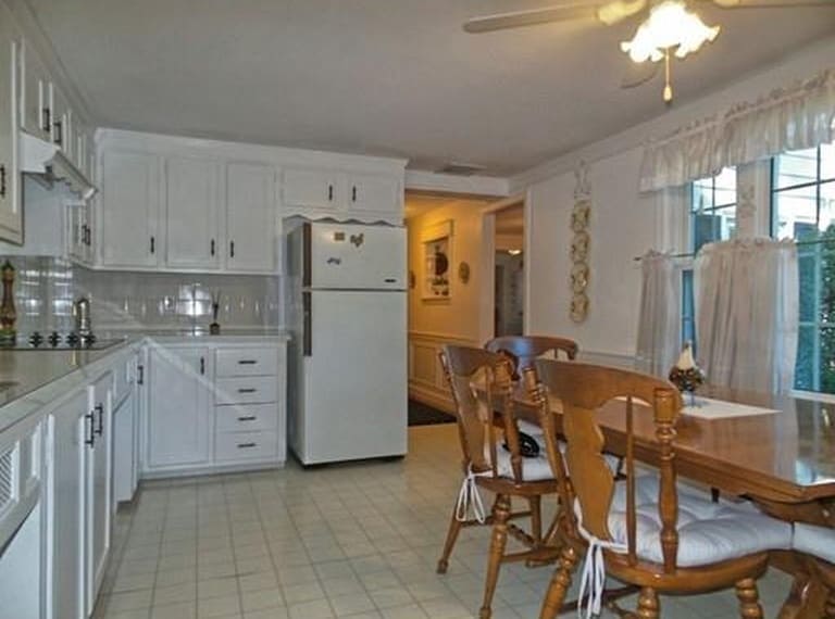 A kitchen with white cabinets, white refrigerator, white tile flooring and a wooden table and country-style wooden chairs with tied-on cushion seats
