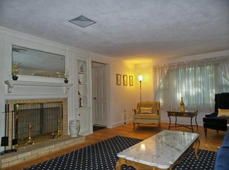 A living room with a fireplace, arm chair & lamp in corner near a window and a large, rectangular coffee table and blue area rug