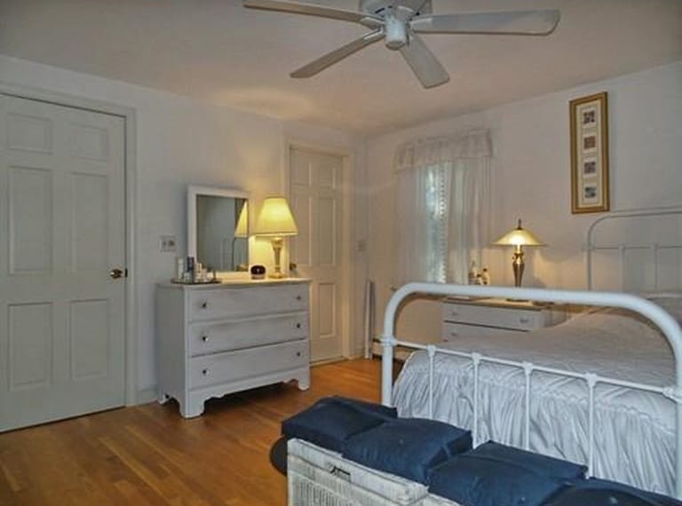 A bedroom with a ceiling fan, an old-fashioned iron bed, a chest at the end of the bed with blue pillows and a dresser with a lamp and mirror