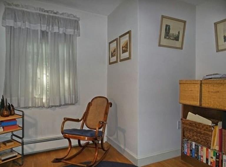 A wooden rocking chair near a window with white curtains, older frames on the wall, and small bookshelves nearby